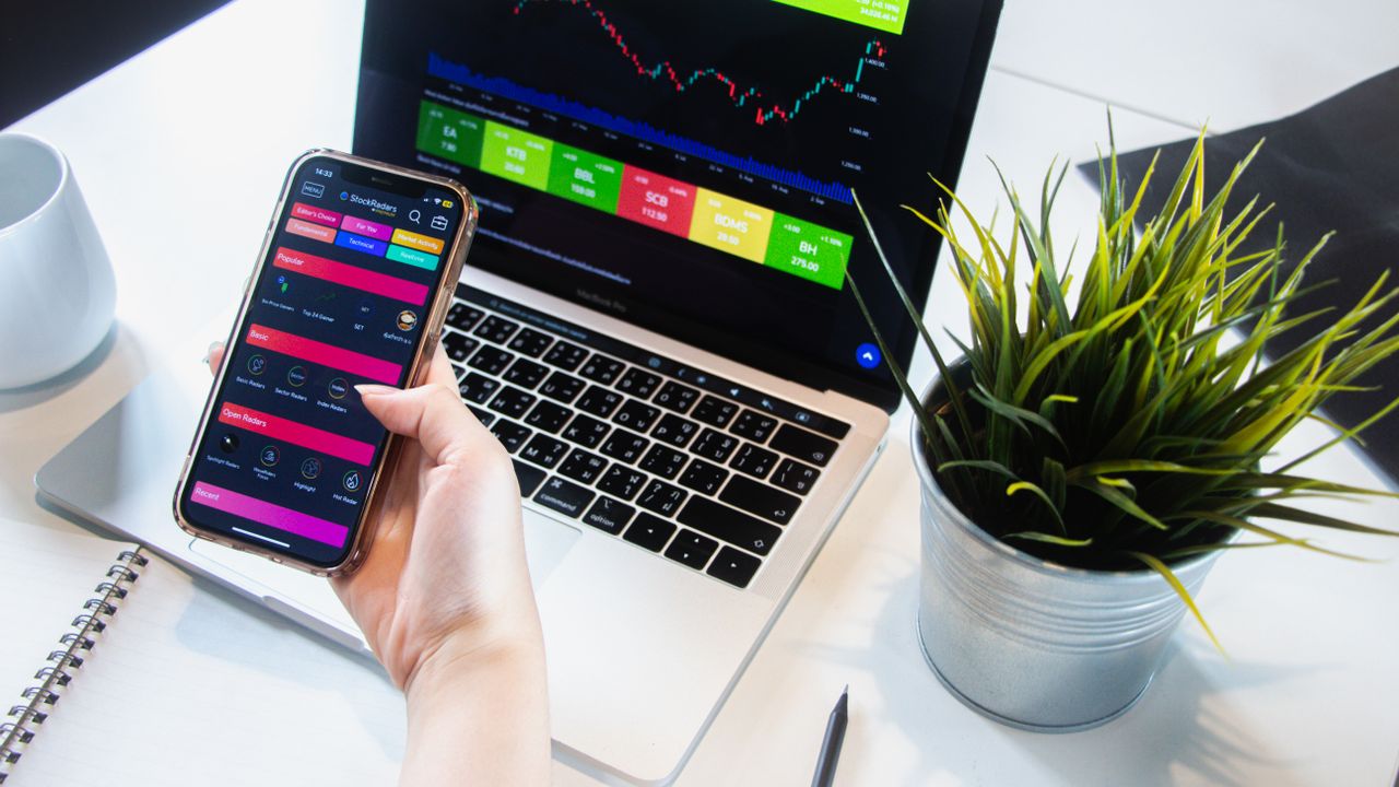 process-img A person using a smartphone app to trade stocks with a laptop displaying market data.