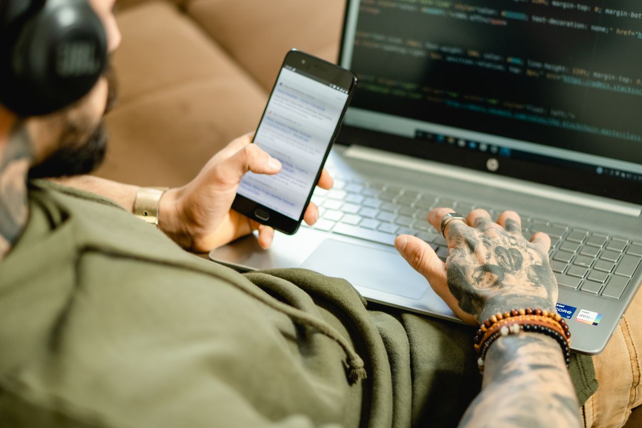 about-01 A tattooed person engaged in coding on a laptop while checking a smartphone.